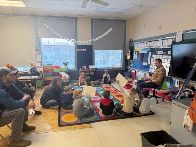 Elementary classroom circle time with a teacher seated at the front and young students sitting on a colorful rug, some holding up papers, while a few adults observe from chairs along the wall.