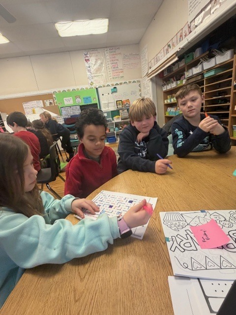 Four upper elementary students seated around a table working on a paper activity with markers and worksheets, while other groups work in the background.