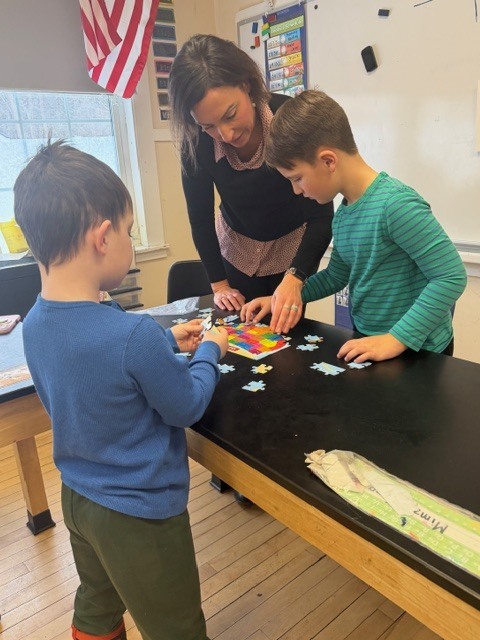 Teacher leaning over a table helping two students assemble a jigsaw puzzle, with puzzle pieces spread across a black tabletop.