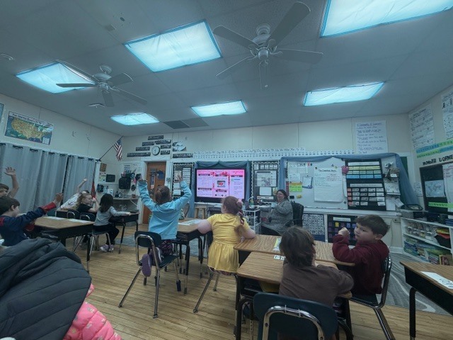 Wide classroom view with students seated at desks raising their hands while a teacher leads a lesson at the front near a large display screen.