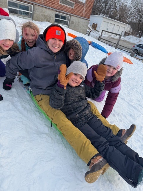 Six children in winter coats, hats, and mittens smile and wave while sitting together on a green sled at the top of a snowy schoolyard hill.
