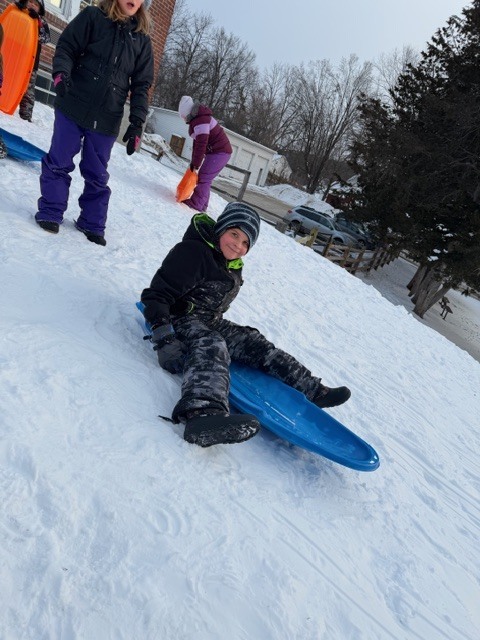 Child in winter gear sits on a blue sled partway down a snowy hill while other students with sleds stand and prepare behind them.