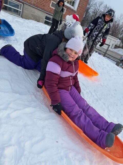 Two children share an orange sled on the snow, one seated and smiling at the camera while another leans forward behind them, with more students and sleds nearby.