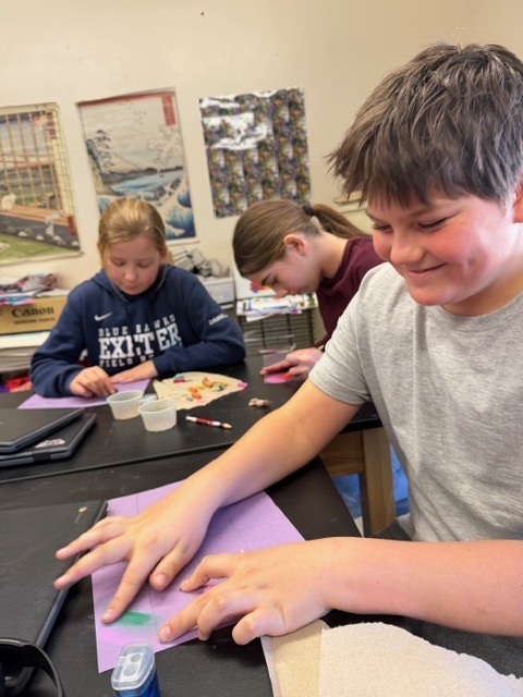 Older students at an art table shaping and pressing small colorful pieces of modeling material onto purple paper while working on a mixed-media project.