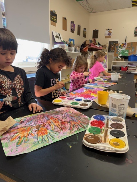 Students lined up at an art table carefully painting bright, layered handprint artwork, with paint palettes and water cups spread across the surface.