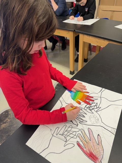 Student in a red shirt coloring a handprint-themed worksheet with a pencil while classmates work quietly at tables in the background.