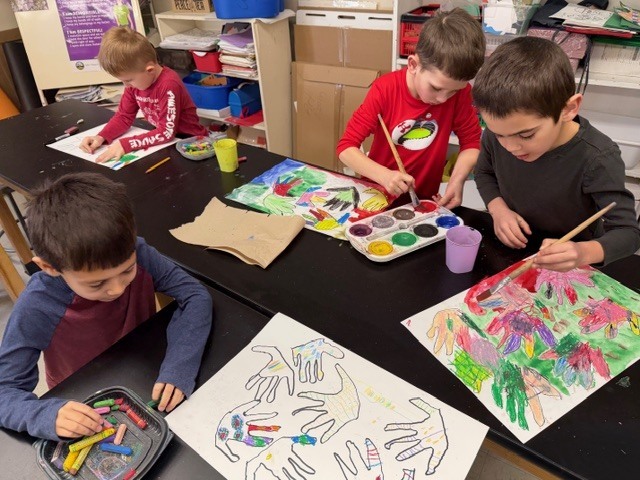 Group of elementary students working at a table, some coloring handprint drawings with oil pastels while others paint with watercolors on similar hand-themed art projects.