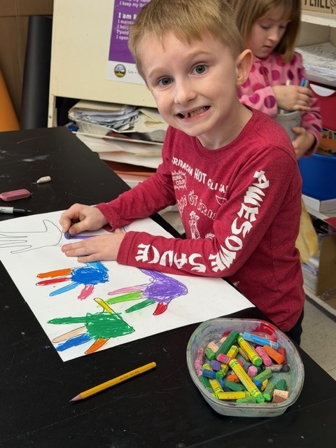 Smiling student seated at a classroom table coloring a handprint art page with oil pastels, with a container of pastels and pencils nearby.