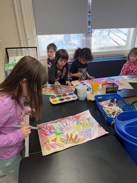 Several young students stand around a classroom art table painting colorful handprint-style designs with watercolor paints and brushes near a window.