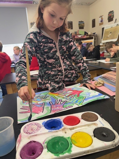 Student painting a multicolored handprint artwork with a brush and watercolor palette in the foreground while classmates work at tables behind.