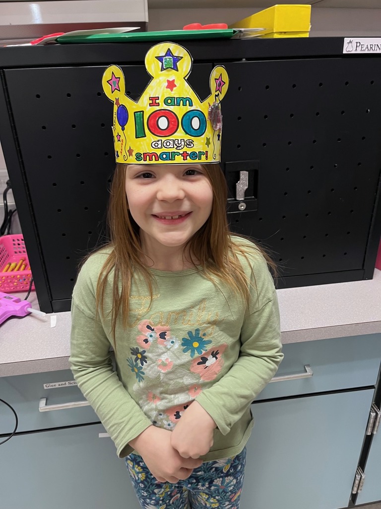  A young girl with long brown hair smiles in front of a black cabinet. Her crown is colored entirely in bright yellow marker, making the "100" stand out in bold green, red, and blue.  i