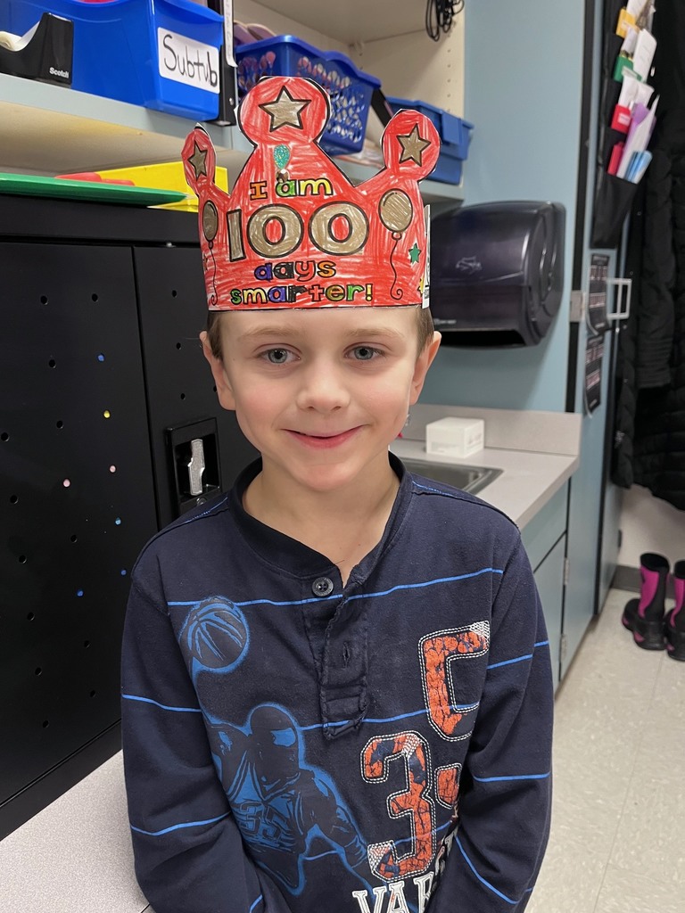 A boy with light blue eyes and a subtle smile wearing a crown colored in vibrant red. He is wearing a dark blue long-sleeve shirt with a basketball graphic.