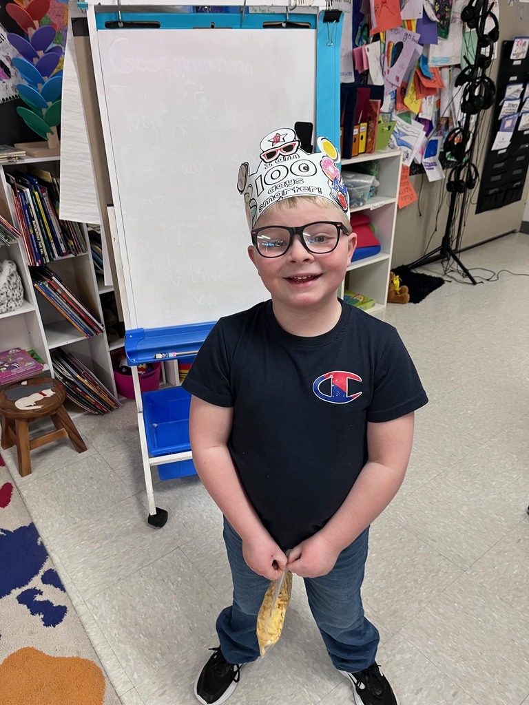 A boy with glasses wearing a navy blue Champion t-shirt, standing in a classroom and holding a small plastic bag of snacks. His crown features a sticker of a dog wearing sunglasses.