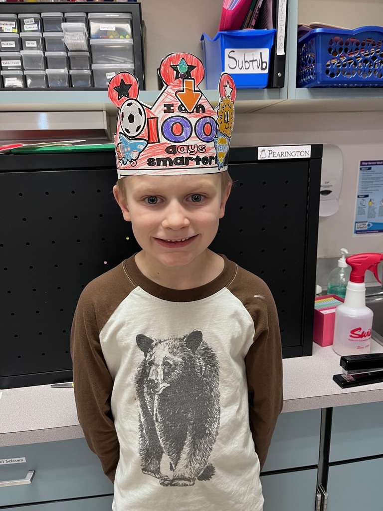  A boy wearing a t-shirt with a large bear graphic stands in a classroom. His crown is heavily decorated with red marker and features a soccer ball sticker and a sun sticker on the sides.  