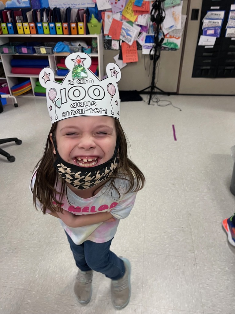 A young girl leans forward with a giant, toothy grin, her face partially framed by a patterned face mask tucked under her chin. She wears a white paper crown decorated with pink balloons and a green glittery tree that says "I am 100 days smarter!"