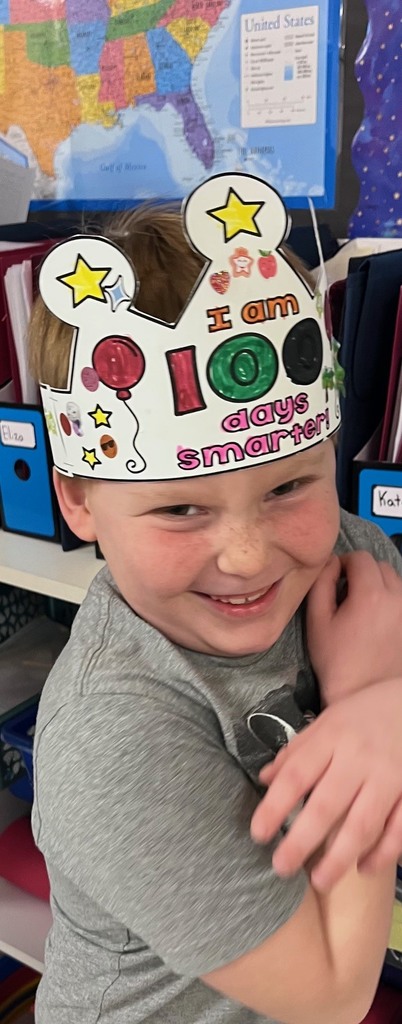 A close-up of a smiling boy wearing a brightly colored crown. He has used markers to color the "100" in pink and green and added star stickers to the points of the crown. A map of the United States is visible in the background.