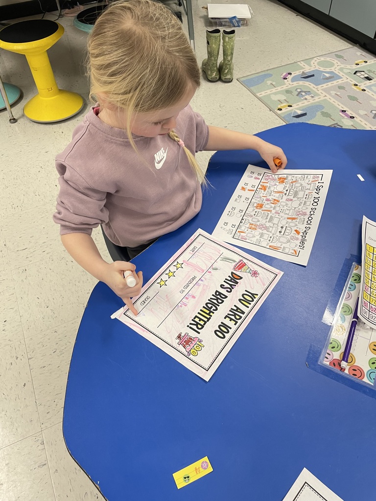 A young girl in a lavender sweatshirt works on two documents: her school supply "I Spy" sheet and a commemorative certificate. She appears to be signing her name at the bottom of the certificate.