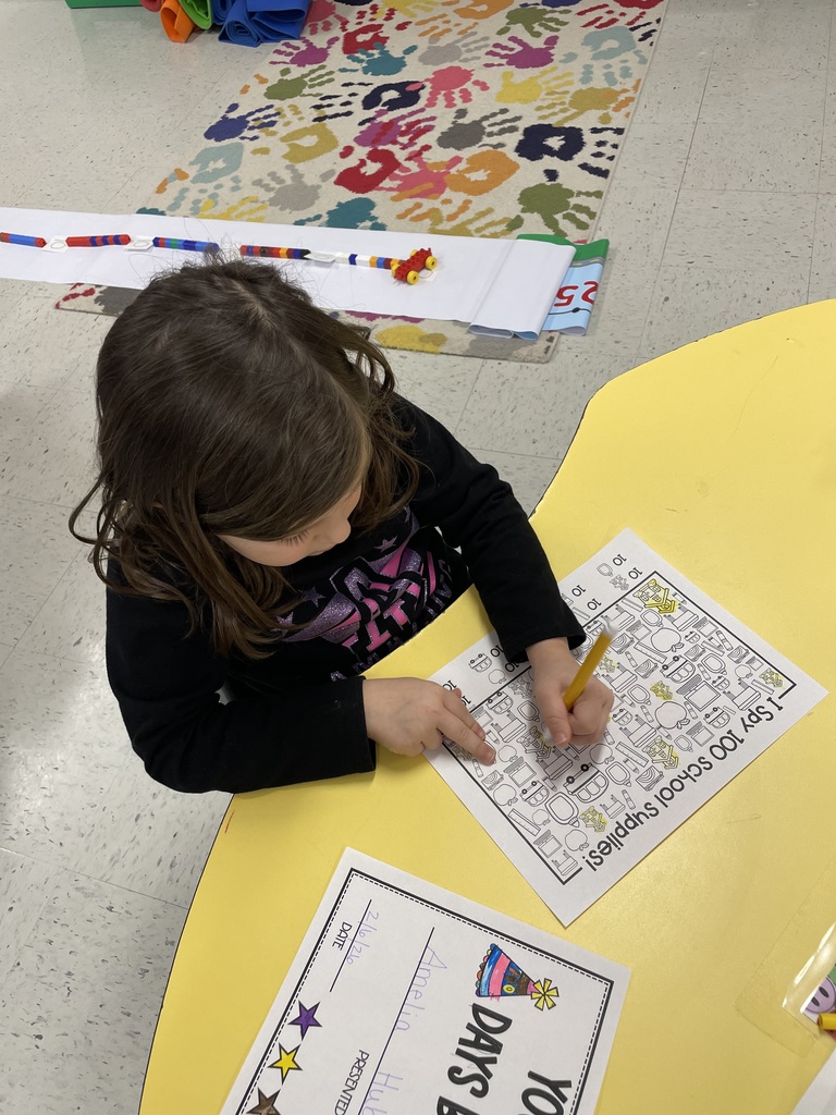 A top-down view of a student at a yellow table working on an "I Spy 100 School Supplies!" worksheet. In the background, a long paper number line is being constructed on the floor using colorful plastic interlocking blocks.