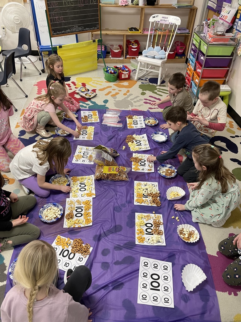 A group of young students sits in a circle on a colorful classroom rug, working on a snack-sorting activity. They are placing different treats—like pretzels, popcorn, and crackers—onto paper templates featuring ten circles to count out exactly 100 items.