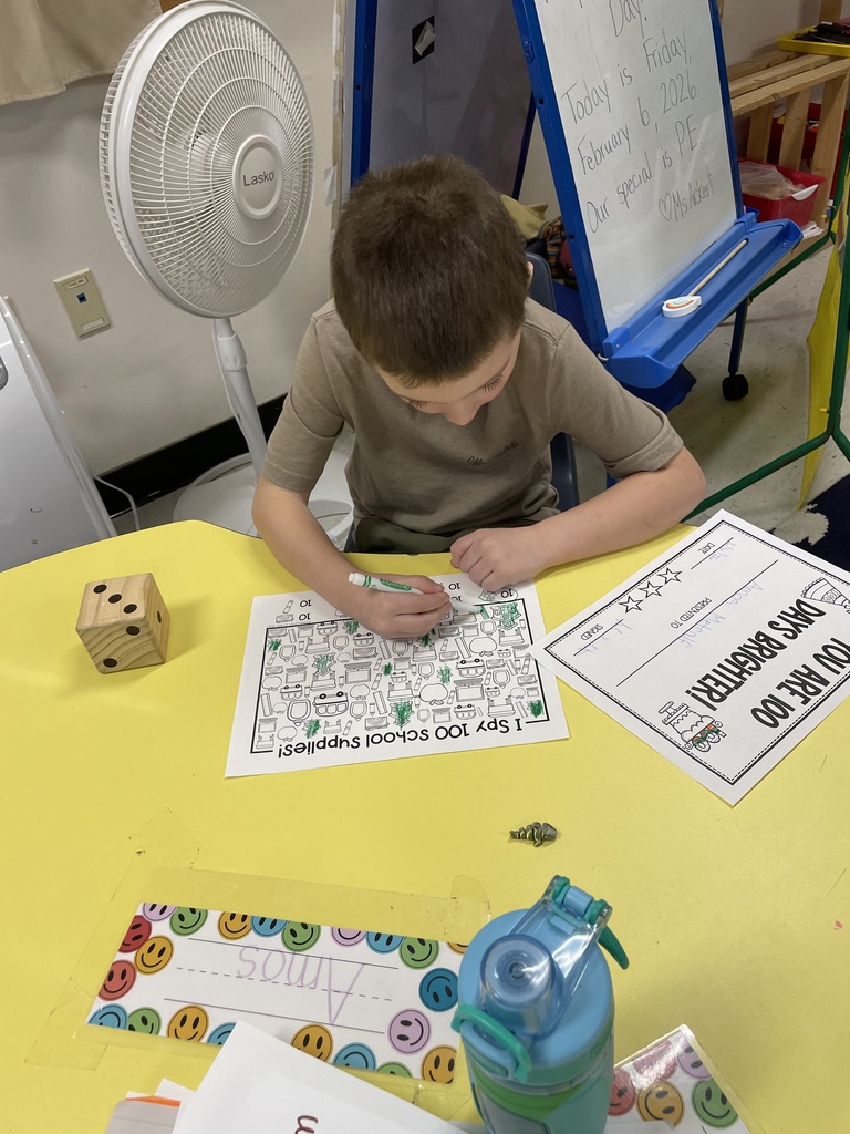 A boy in a tan t-shirt uses a green marker to find and color items on his 100th-day worksheet. A whiteboard in the background displays the date: "Friday, February 6, 2026".