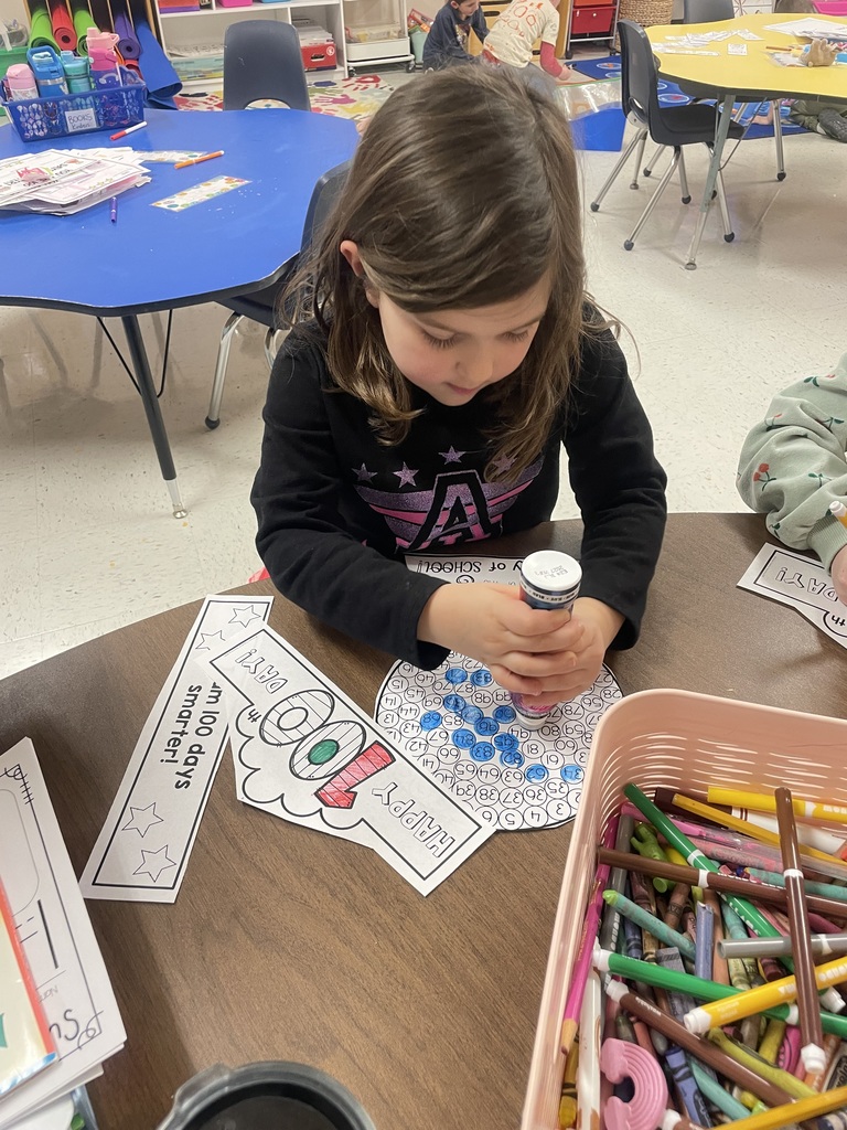 A young girl in a black shirt uses a blue dot marker to fill in circles on a "100th Day" activity sheet. Beside her is a large bin overflowing with colorful markers and crayons.