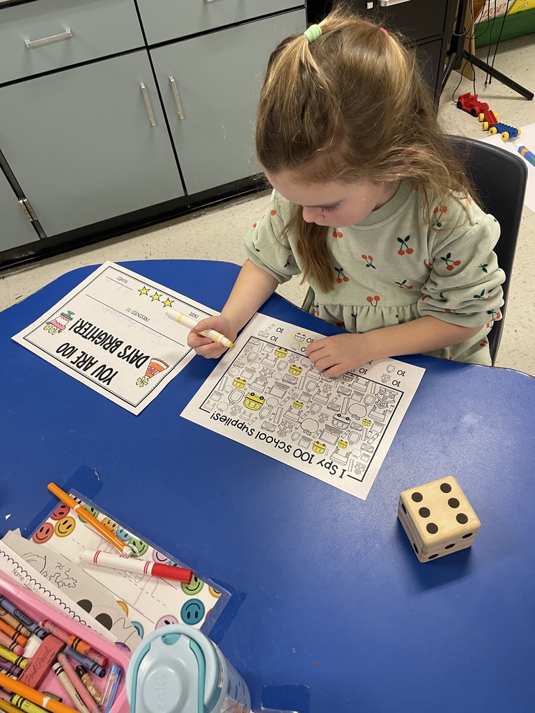 A student in a light green floral dress focuses on coloring small school supply icons on her worksheet. A large wooden die and a container of crayons sit on the blue table in front of her.