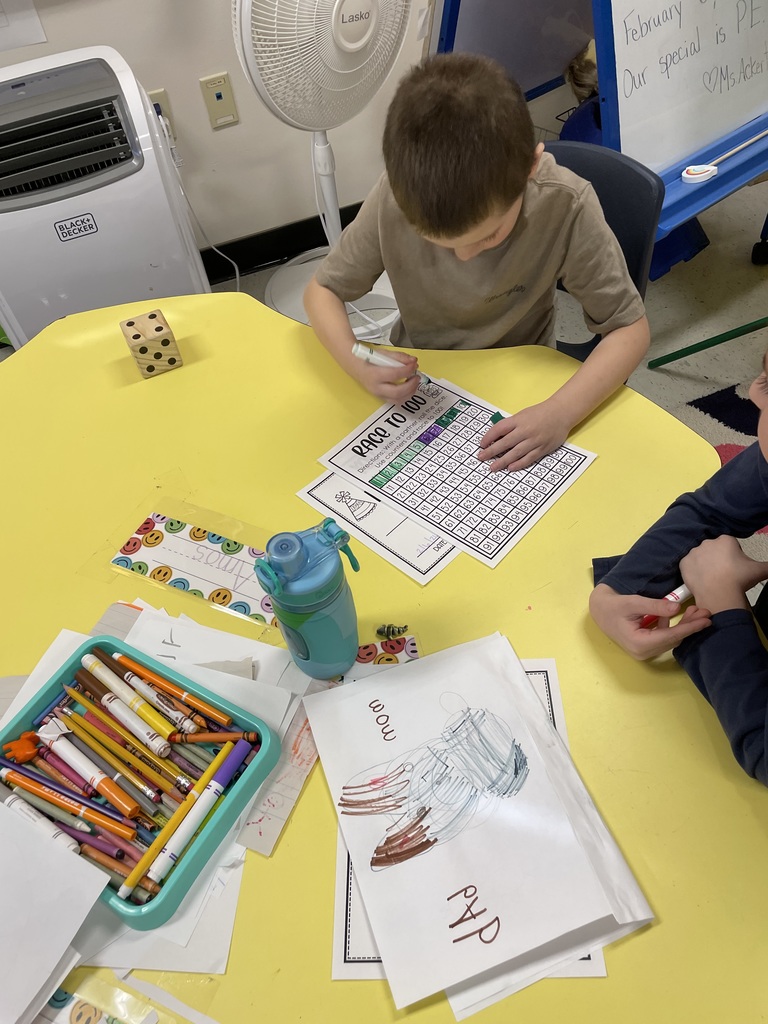 A student at a yellow table uses a green marker to find hidden items on a worksheet. A classroom whiteboard in the background shows the date as Friday, February 6, 2026.