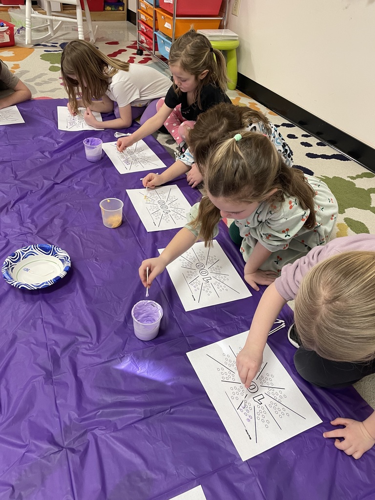 A group of students sits on a colorful handprint-patterned rug, participating in a snack-sorting activity. They are carefully placing small snacks like pretzels, popcorn, and goldfish crackers into ten printed circles on "100" templates to reach a total of 100 treats.