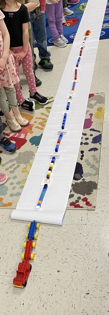 A group of young children stand around a long white paper strip laid out on a classroom floor, with colorful LEGO trains and blocks arranged in a straight line down the center. Only the children’s legs and shoes are visible as they watch, and a patterned classroom rug borders part of the scene.”