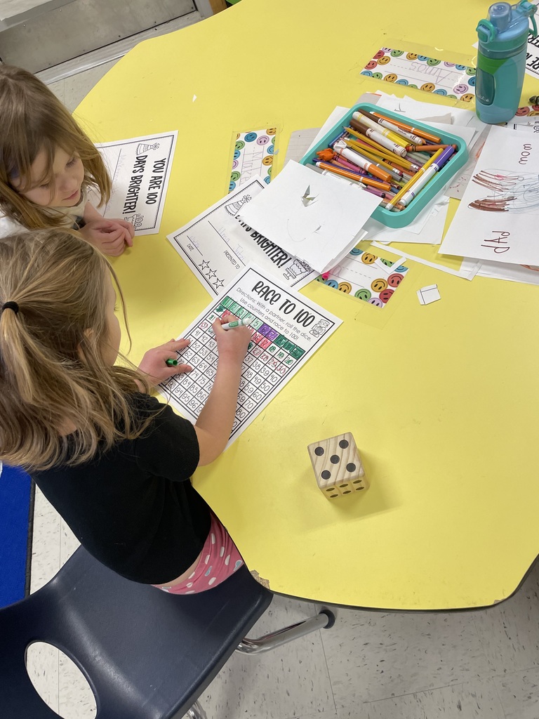 “Two young children sit at a round yellow classroom table working on a ‘Race to 100’ math activity. One child colors in numbers on a hundreds chart with a green marker while the other watches. A large wooden die, a tray of crayons and markers, worksheets, and a water bottle are spread across the table.”