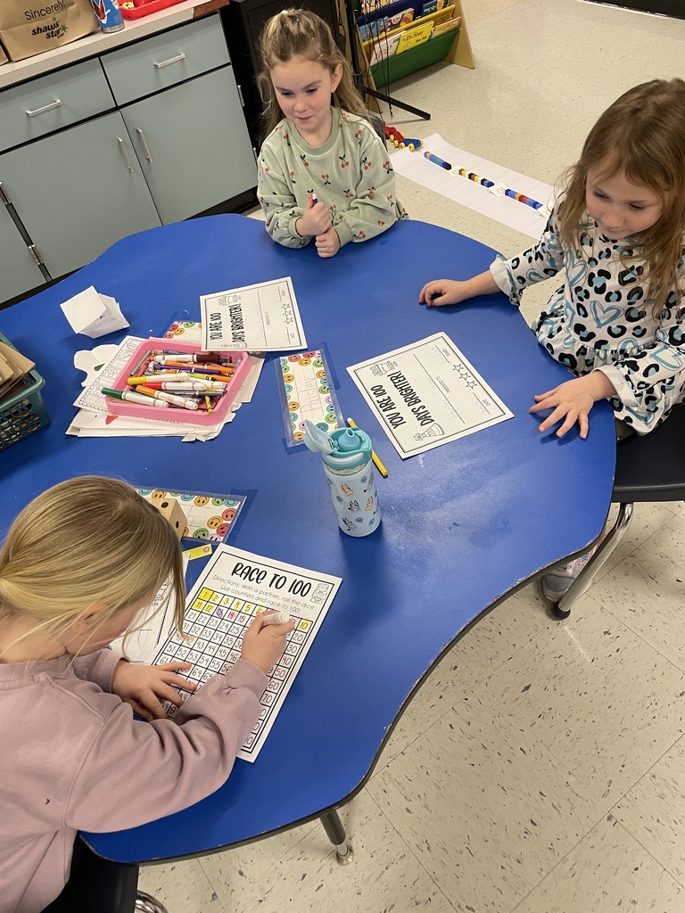 Three young children sit around a blue classroom table working on ‘Race to 100’ math worksheets. One child colors in numbers on a hundreds chart while the others watch and wait their turn. Crayons, markers, papers, and a water bottle are on the table, and a long line of colorful blocks on paper is visible on the classroom floor in the background.”