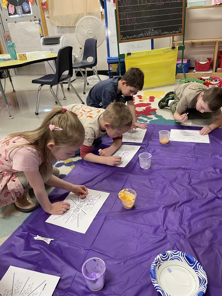A group of students sits on a colorful handprint-patterned rug, participating in a snack-sorting activity. They are carefully placing small snacks like pretzels, popcorn, and goldfish crackers into ten printed circles on "100" templates to reach a total of 100 treats.