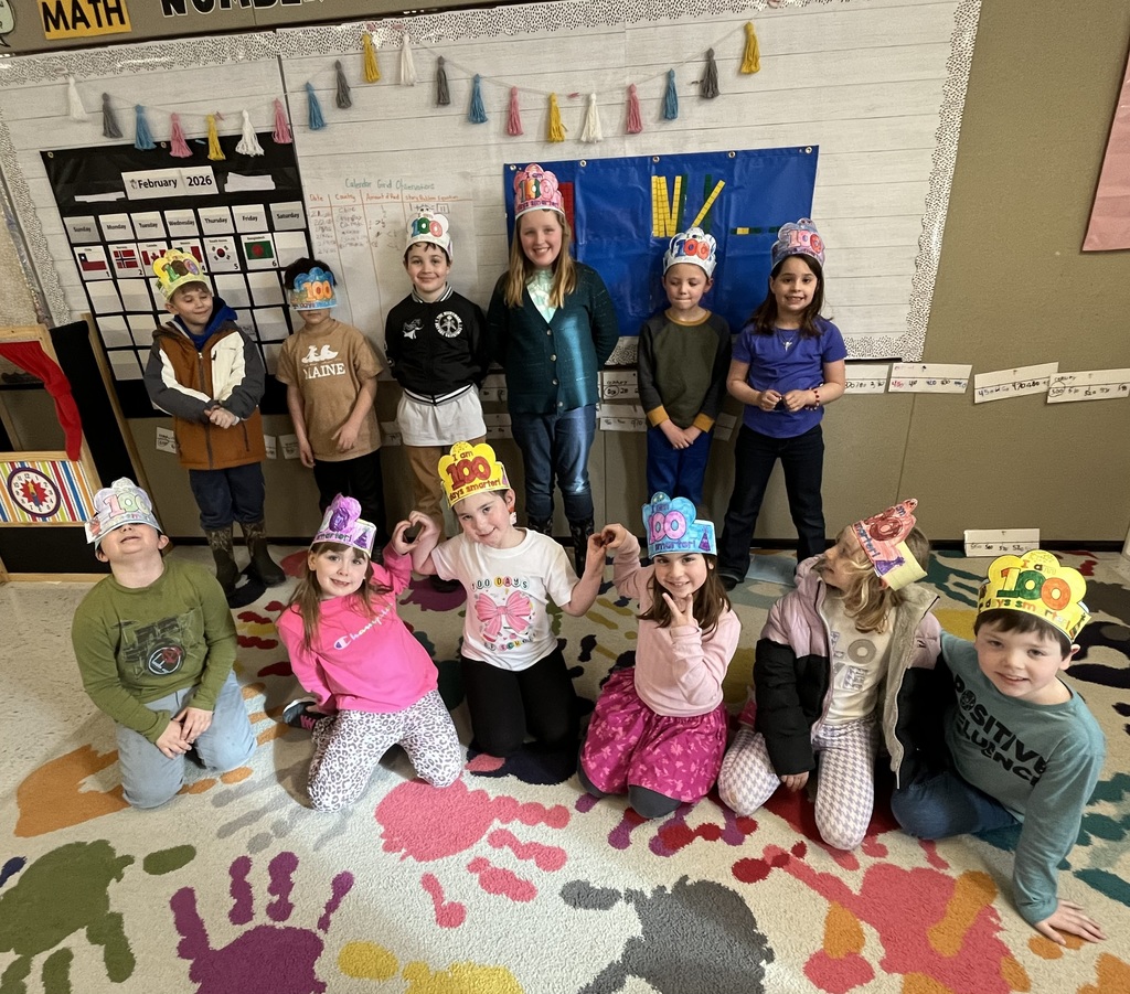 A group of twelve young students and their teacher pose in a classroom, celebrating the 100th day of school. All the children wear colorful, handmade paper crowns that say "100" or "100 Days Smarter." Six children are standing in the back row with their teacher, and six are kneeling or sitting on a vibrant rug decorated with colorful handprints. The classroom background features a "MATH" sign, a February 2026 calendar, and a blue pocket chart displaying yellow counting sticks.