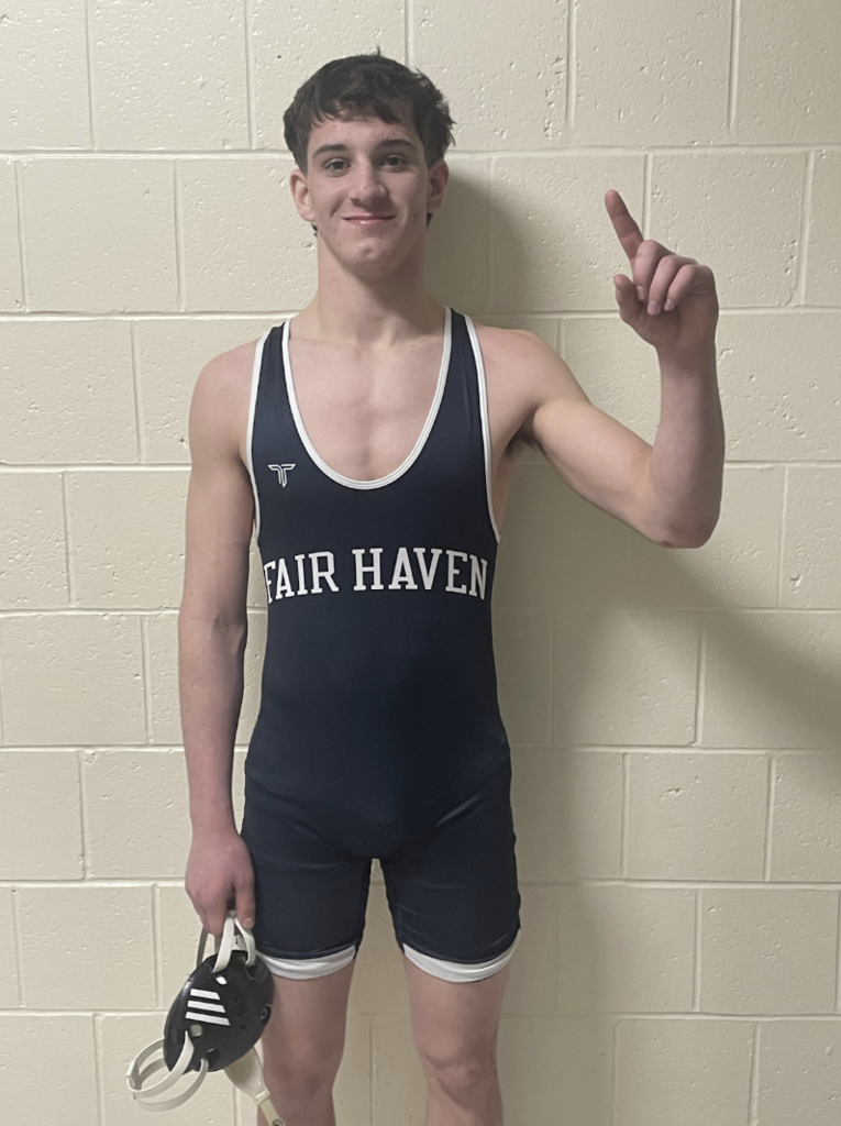 Student wrestler in a navy Fair Haven singlet stands against a cinderblock wall, smiling and holding up one finger with his right hand while holding his wrestling headgear in his left.
