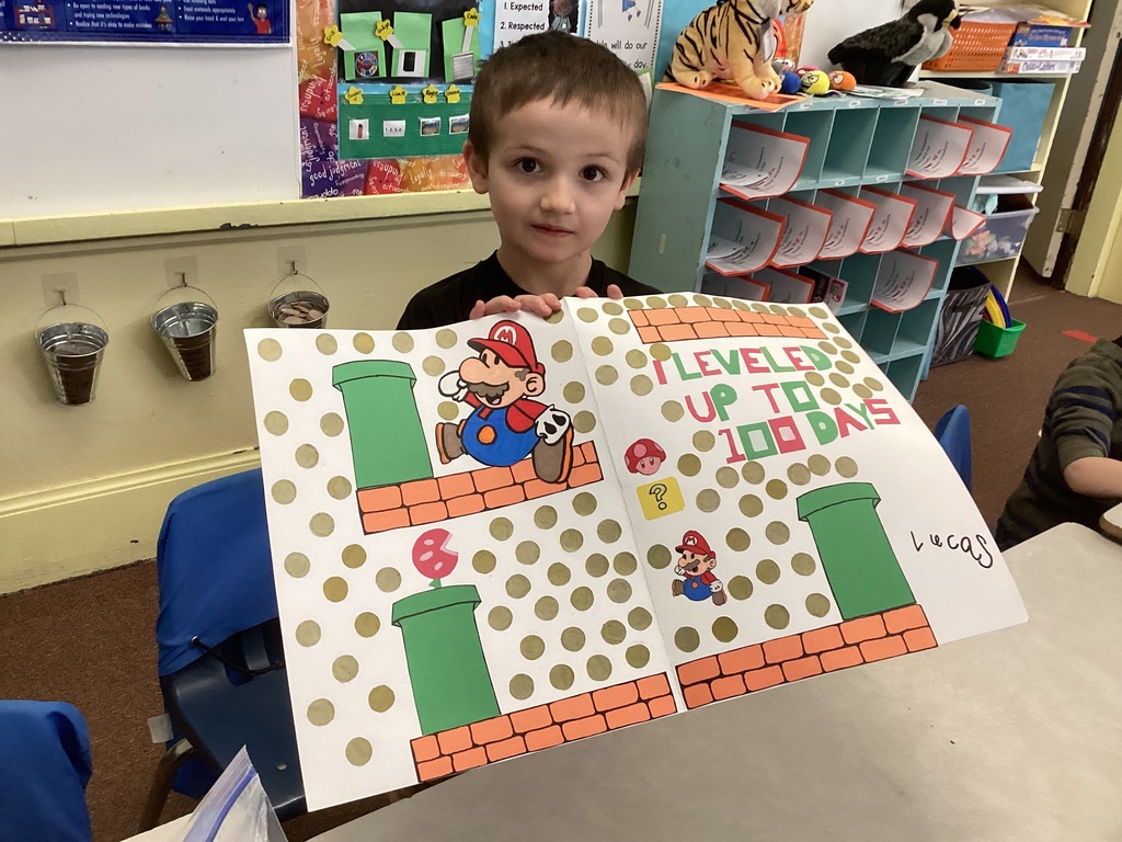 Student sitting at a classroom table holding a handmade poster with a Super Mario theme that reads “I leveled up to 100 days,” decorated with gold dots and green pipes.