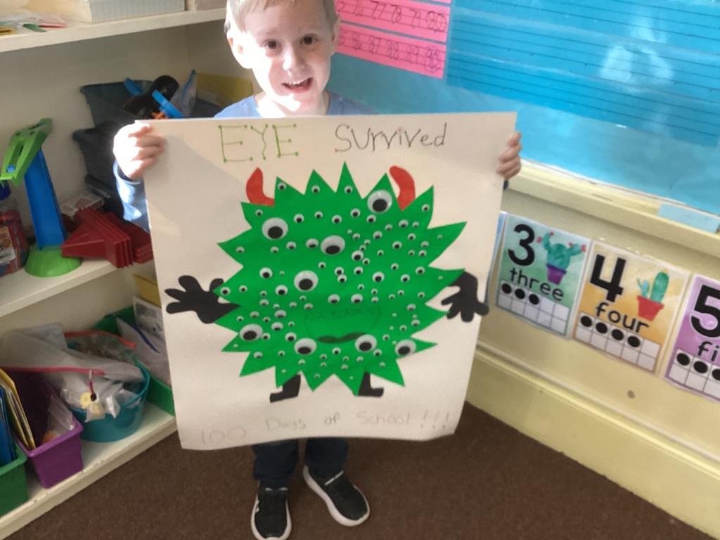 Student standing in a classroom holding a large green monster-themed poster with many googly eyes that says “I survived 100 days of school.”
