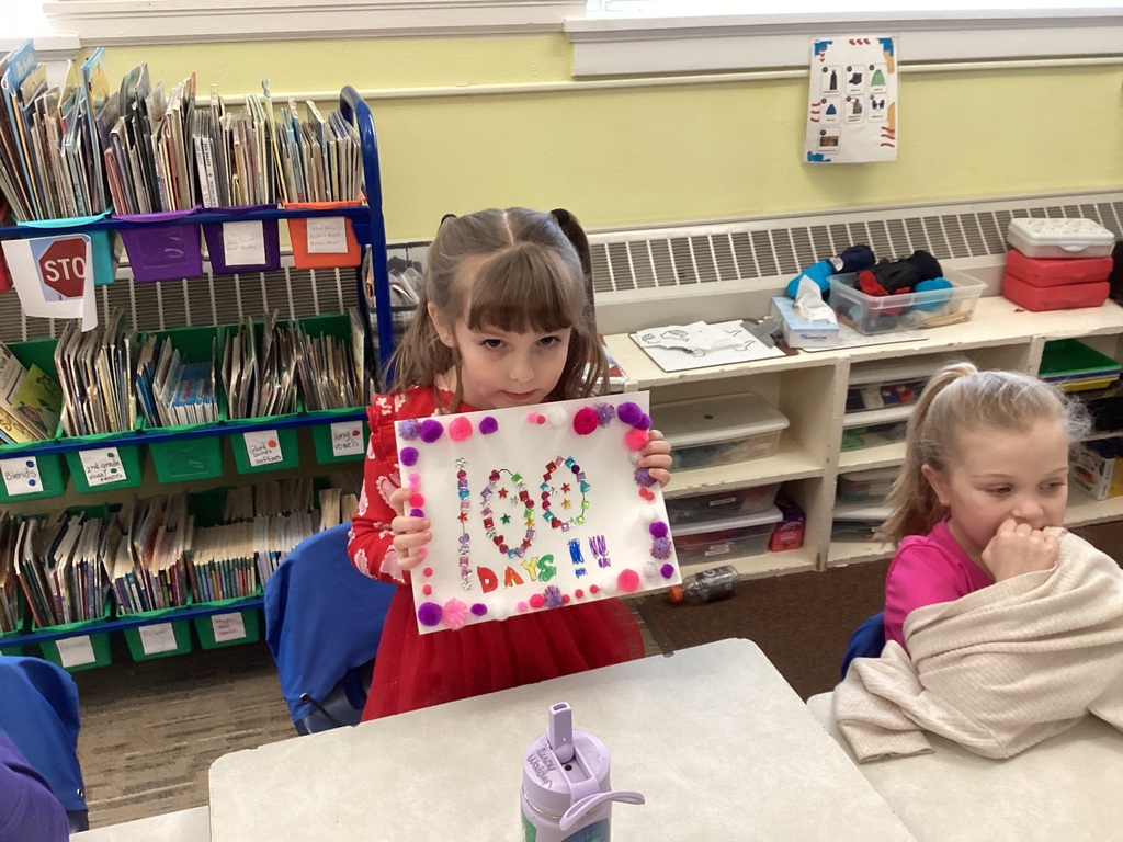 Young student in a classroom holding a decorated “100 days” project board covered with colorful gems and pom-poms, with bookshelves and bins in the background.