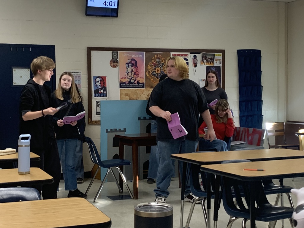 High school students stand at the front of a classroom holding scripts and performing a scene, with desks in the foreground and posters and a bulletin board on the wall behind them.