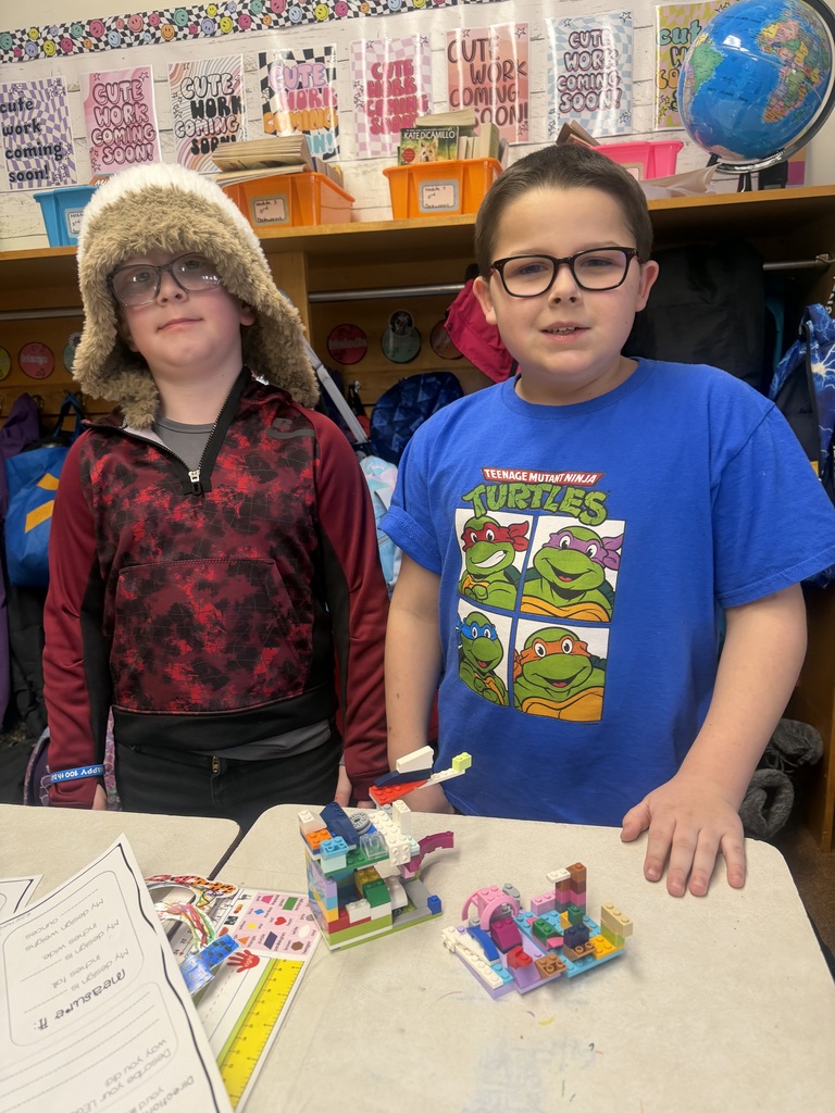Two students standing at desks with their LEGO builds and challenge papers, posing in a classroom with cubbies and school supplies visible behind them.