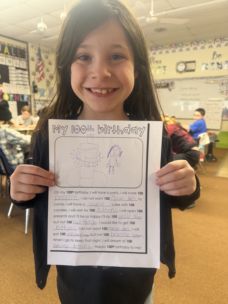 Student smiling and holding a worksheet titled “My 100th birthday” with a drawing and writing about what they would do on their 100th birthday.