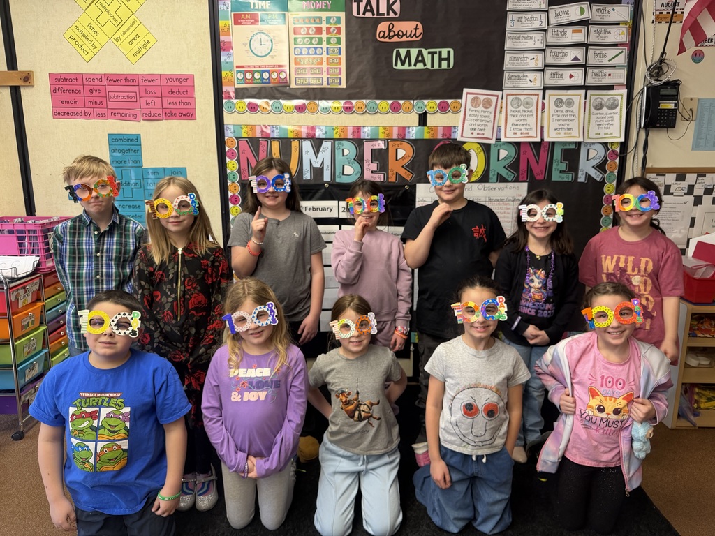Group of elementary students standing in a classroom in front of a “Number Corner” math bulletin board, smiling and wearing colorful handmade “100 days” glasses to celebrate the 100th day of school.