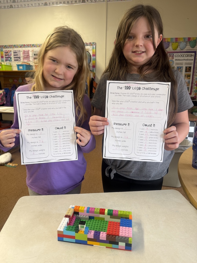 Two students smiling and holding “100 LEGO Challenge” worksheets while standing beside a tall, multicolored LEGO build on a classroom desk.