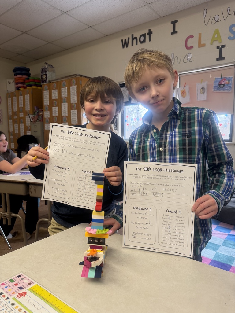 Two students standing behind a desk holding completed “100 LEGO Challenge” worksheets, with a colorful LEGO structure displayed in front of them.