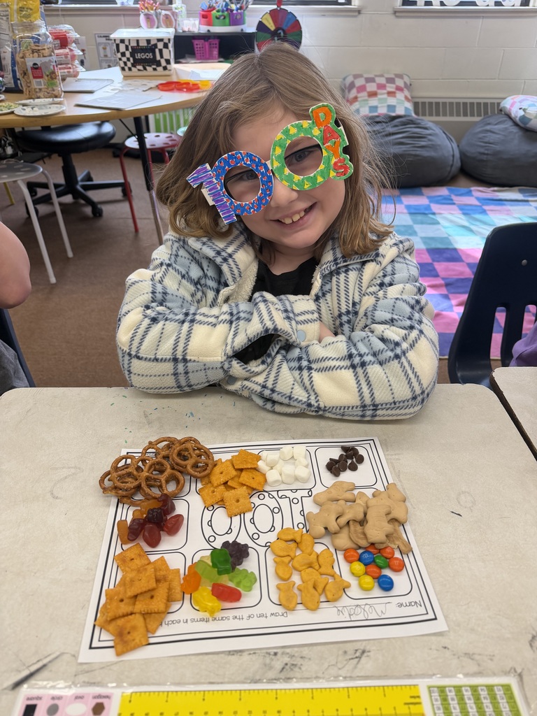 Student wearing decorated “100 days” glasses sits at a desk with a worksheet and an assortment of snacks sorted into groups for a 100th day counting activity.