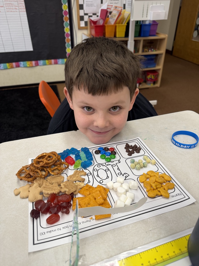 Student smiling at a desk with a “100 days” snack mat, showing grouped treats like pretzels, crackers, marshmallows, and candy arranged for a 100th day math activity.