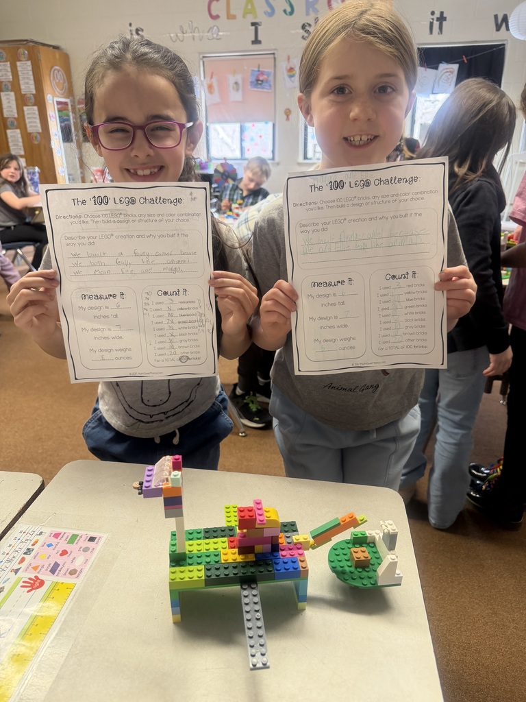 Two students standing behind a desk holding completed “100 LEGO Challenge” worksheets, with a colorful LEGO structure displayed in front of them.