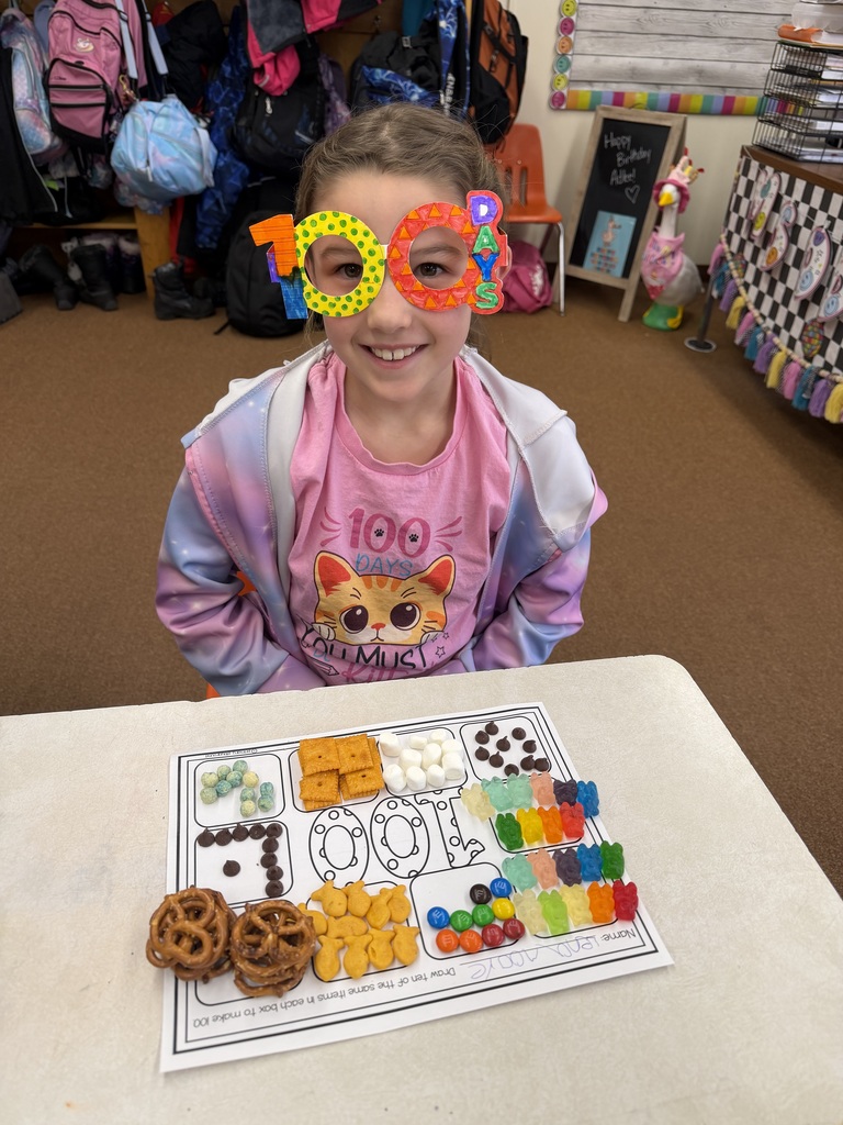 Student wearing bright “100 days” glasses sits at a desk with a worksheet and neatly sorted snacks and candies arranged in groups to total 100 items.