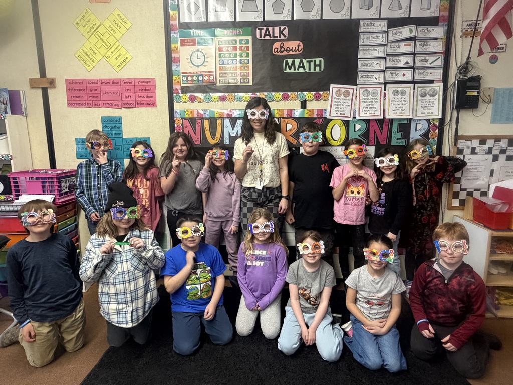 Class photo of students and a teacher in front of a math bulletin board, many wearing decorated “100 days” glasses and posing together for a 100th day of school celebration.