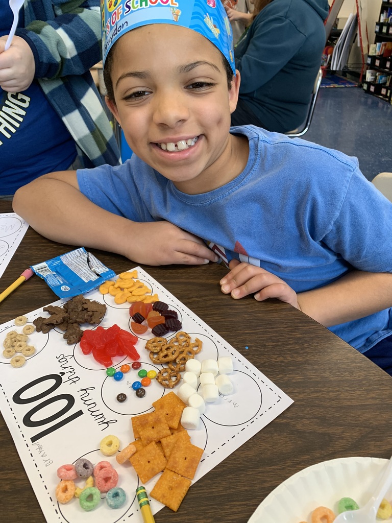 Smiling third grade student wearing a “100 Days of School” crown sits at a desk with a worksheet and a snack display arranged into groups of 100 using cereal, crackers, candy, pretzels, and marshmallows.