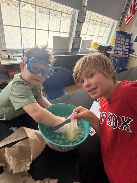 Two students in goggles lean over a teal bowl of ice water while holding a plastic bag, smiling during a hands-on experiment.